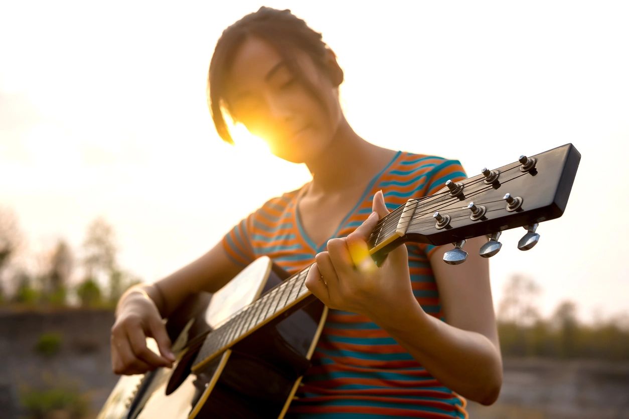 woman playing acoustic guitar with sun behind her - electric vs. acoustic guitar 